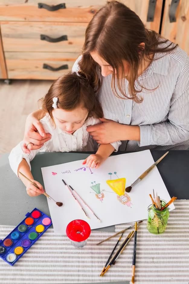 Mulher acompanha uma menina pintando em uma mesa